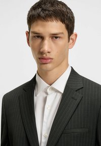 Black pinstriped suit jacket with notch lapels, paired with a white collared shirt. Smooth fabric with a tailored fit.