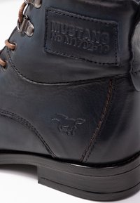 Černá kožená bota s hnědými tkaničkami, vybavená kovovými průvleky. Na boku je vyznačeno značkové logo a embosovaný detail koně. Plná černá podrážka.