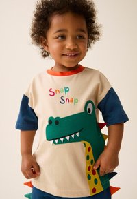 Cotton t-shirt with short blue sleeves, cream body, green dinosaur graphic, colorful lettering "Snap Snap," and triangle accents on the side.