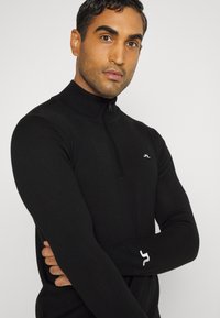 Black zip-up sweater with a high collar, made of textured fabric. Features a small logo on the chest and a white accent on the sleeve.