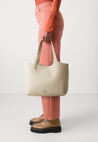 Beige-värinen kangaskassi, jossa on sileä pinta ja yksinkertainen olkahihna, sekä hillitty logo. Yhdistetty vaaleanpunaisten housujen ja vaaleanruskeiden kenkien kanssa.