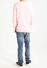 Person, der mit dem Rücken zur Kamera steht, trägt einen hellrosa Pullover, blaue Jeans und schwarze Sneakers vor einem weißen Hintergrund.