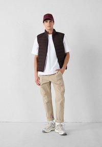 Donkerbruine puffer-vest over een wit t-shirt, gecombineerd met beige cargobroek. Witte en grijze sneakers, bordeauxrode pet, minimalistisch design.