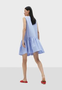 Light blue striped sleeveless dress with a flared skirt, featuring a gathered hem. Paired with red slip-on shoes. Smooth fabric texture.