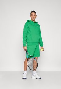Conjunto de hoodie verde e calções, material de algodão, com riscas laterais brancas. Asegurando uma raquete de ténis e uma bola, ténis brancos.