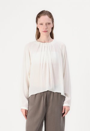 Hvit bluse med lange ermer, plissert front, rund hals og flytende silhuett. Kombinert med grå, løstsittende bukser.