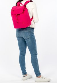 Růžový batoh s skládacími klopami, nastavitelnými popruhy a logem. Nosený s modrými džíny a bílými teniskami.