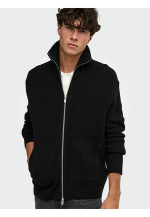 Jeune homme aux cheveux bouclés porte un pull noir côtelé zippé sur une chemise blanche, mains dans les poches, sur un fond clair uni.