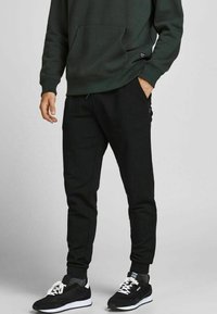 Zwarte joggingbroeken met opgerolde enkels, gemaakt van een glad materiaal. Gepaard met een donkergroen sweatshirt en zwarte sportschoenen.