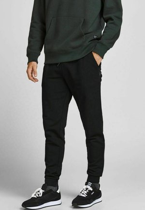 Zwarte joggingbroeken met opgerolde enkels, gemaakt van een glad materiaal. Gepaard met een donkergroen sweatshirt en zwarte sportschoenen.
