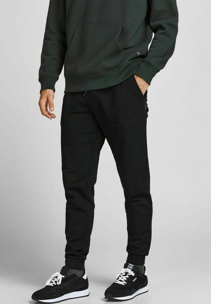 Zwarte joggingbroeken met opgerolde enkels, gemaakt van een glad materiaal. Gepaard met een donkergroen sweatshirt en zwarte sportschoenen.