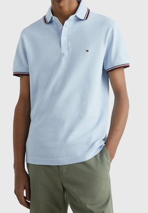 Man draagt lichtblauw poloshirt met rode, witte en zwarte bies aan de kraag en mouwen, olijfgroene broek, één hand in zijn zak.