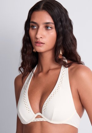 Femme aux cheveux foncés ondulés portant un haut de bikini blanc à col halter orné de petits clous dorés, regardant légèrement sur le côté.