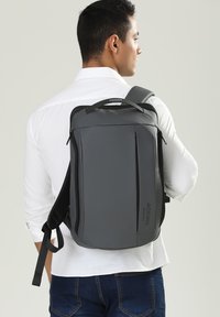 Zaino grigio dal design elegante, con una maniglia superiore, spallacci regolabili, tasche frontali con cerniera e una finitura liscia e opaca.