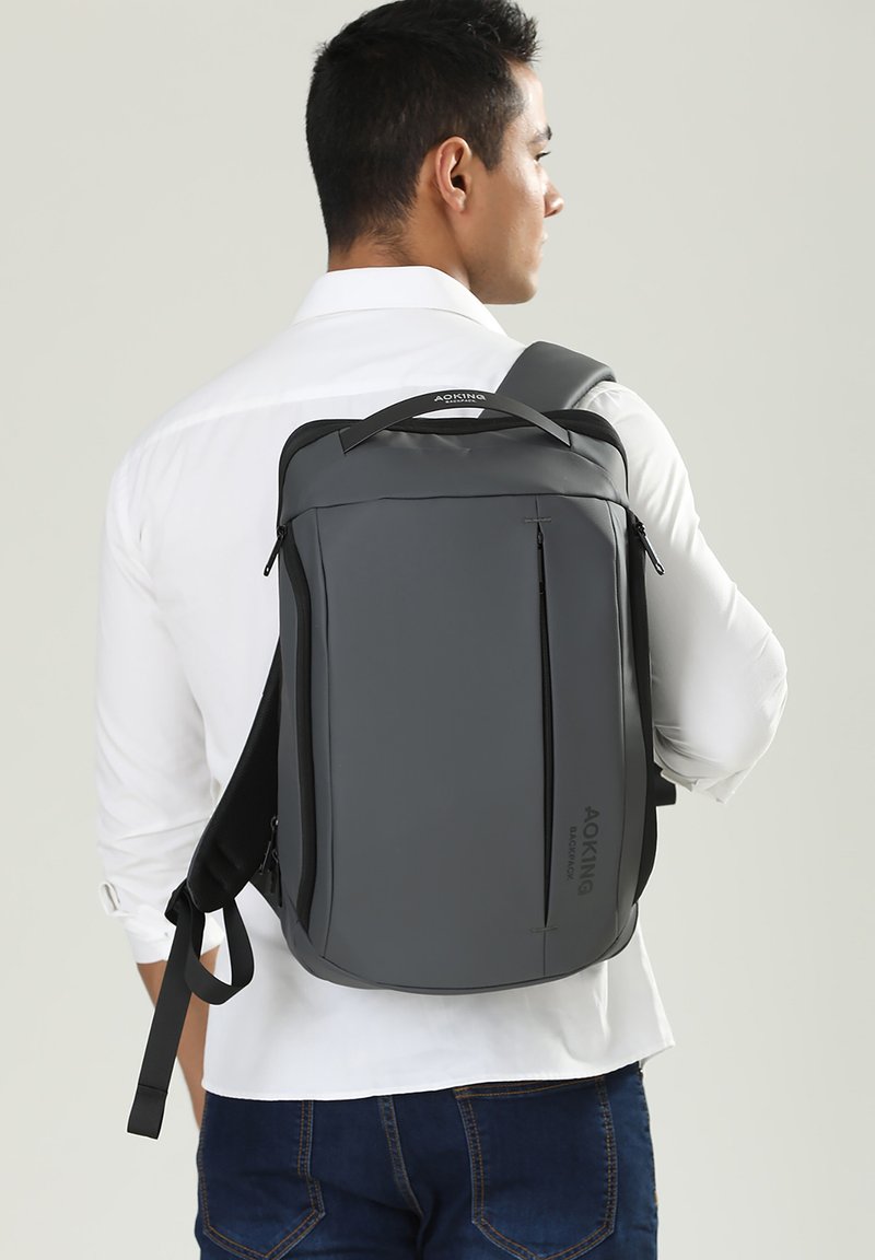Zaino grigio dal design elegante, con una maniglia superiore, spallacci regolabili, tasche frontali con cerniera e una finitura liscia e opaca.