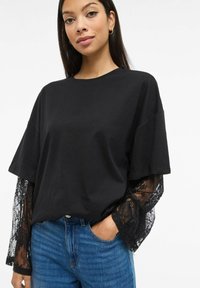 Svart kortermet skjorte med rund halsutringning og gjennomsiktig svart blondedetalj på ermene, kombinert med blå denimjeans.