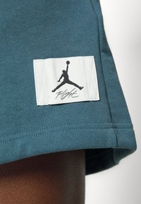 Tissu bleu sarcelle foncé avec un patch rectangulaire blanc montrant une silhouette noire d’un joueur de basket-ball et le mot "Flight" en dessous.