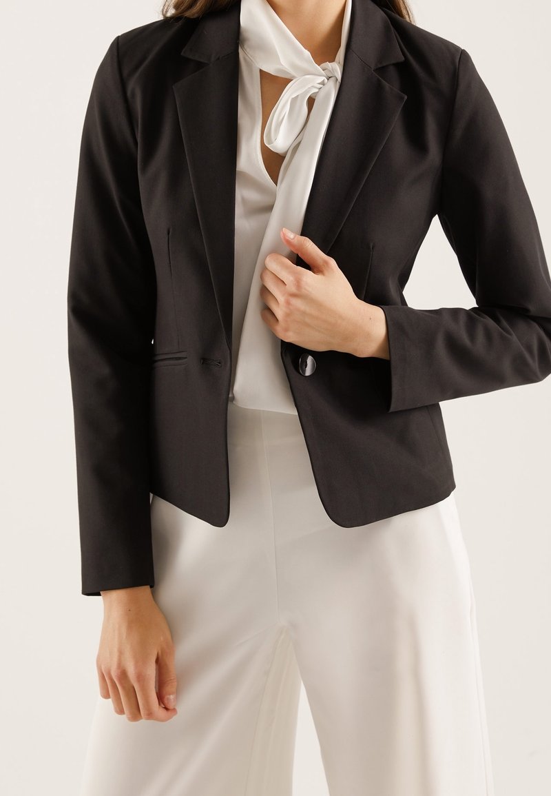 Black blazer with a fitted design, featuring a single button closure, two front pockets, and notch lapels, worn over a white blouse.