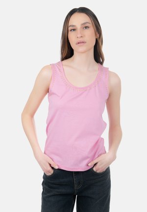 Donna con capelli castani lunghi che indossa una canotta rosa con borchie dorate intorno allo scollo e jeans scuri, con le mani nelle tasche.