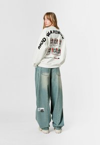 Sudadera blanca de gran tamaño con estampados gráficos en la parte posterior. Combinada con jeans de pierna ancha de un azul desvaído y zapatos de plataforma beige.
