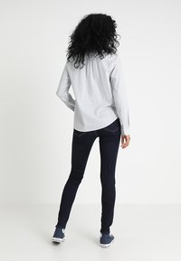 Camicia azzurra chiara a maniche lunghe con colletto, abbinata a jeans skinny scuri e scarpe di tela blu. Lo sfondo è bianco semplice.
