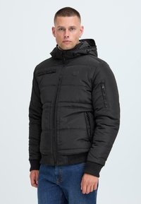 Black puffer jacket with zippered pockets, a hood, and a textured exterior. Features a fitted design with ribbed cuffs and a logo patch.