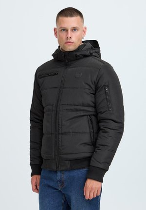 Black puffer jacket with zippered pockets, a hood, and a textured exterior. Features a fitted design with ribbed cuffs and a logo patch.