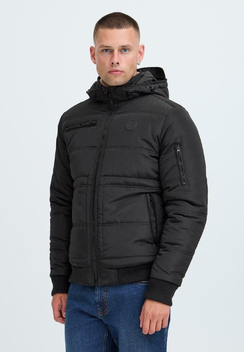 Black puffer jacket with zippered pockets, a hood, and a textured exterior. Features a fitted design with ribbed cuffs and a logo patch.