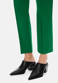 Schwarze Leder-Mules mit spitzer Zehenpartie und klobigem Absatz, kombiniert mit eng anliegenden grünen Hosen im geraden Schnitt und mit klaren Linien.