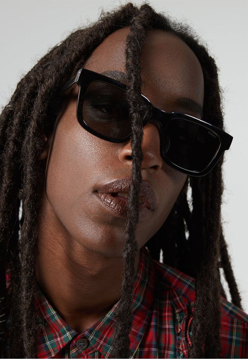 Zwarte vierkante zonnebril met dikke, hoekeigene montuur, gedragen met een geruite overhemd. Lange, gestreepte haren bedekken gedeeltelijk de lens.