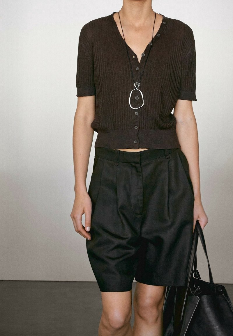 Personne portant un haut côtelé marron foncé à manches courtes avec des boutons, un short noir taille haute, un collier pendentif et tenant un sac noir.