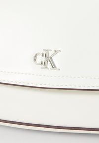 Bijela kožna torba s srebrnim "CK" logotipom i kontrastnim šavovima duž rubova. Glatka tekstura, pravokutni oblik.