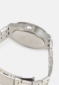 Orologio in acciaio inossidabile con finitura spazzolata, cassa arrotondata e corona metallica circolare. Presenta un bracciale a maglie metalliche e un retro liscio.