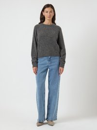 Mulher em pé vestindo um suéter de manga longa cinza escuro, jeans retos de tom azul claro, sapatos bege e brincos de argola dourados em um fundo branco.