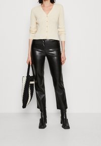 Cárdigan de canalé crema con cuello en V y cierre de botones, acompañado de pantalones de imitación cuero negros de cintura alta. La modelo sostiene un bolso en blanco y negro.