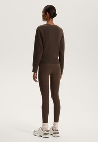 Maglione marrone a maglia con texture a coste, abbinato a leggings marroni. Sneakers grigie e bianche con accenti viola e calzini bianchi al polpaccio.