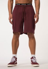 Bermudas de carga granate con cintura elástica, cordón y bolsillos laterales. Fabricadas en una tela suave con un ajuste relajado. Combinadas con zapatillas rojas de caña alta.