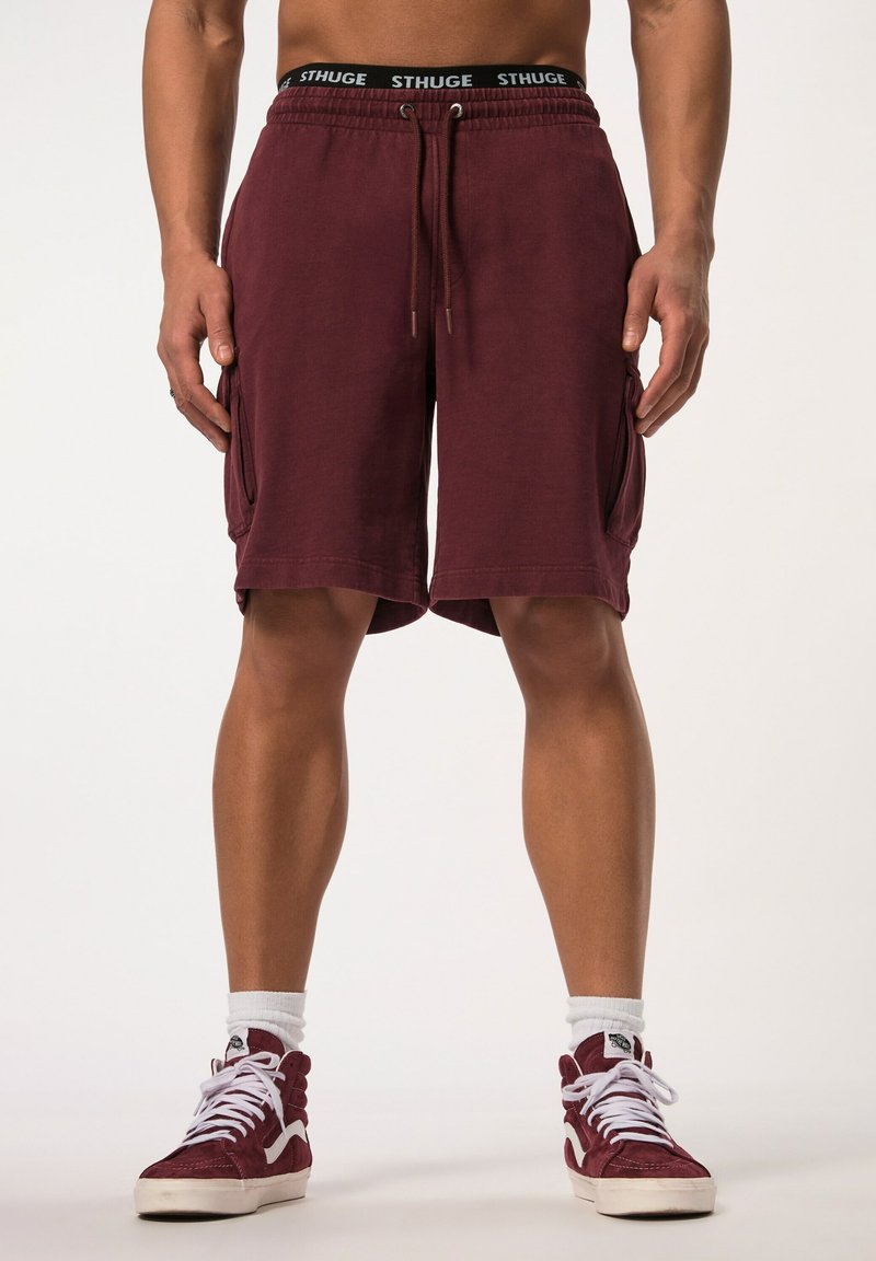 Bermudas de carga granate con cintura elástica, cordón y bolsillos laterales. Fabricadas en una tela suave con un ajuste relajado. Combinadas con zapatillas rojas de caña alta.