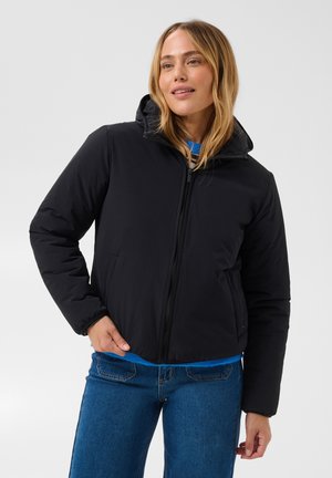 Femme portant une doudoune noire zippée par-dessus un pull bleu et un jean bleu, debout avec une main dans la poche de la veste.
