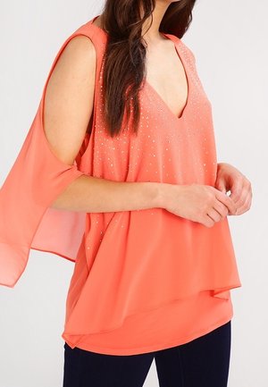 Vrouw draagt een koraalkleurige mouwloze top met V-hals, fonkelende versieringen en open zijkanten, gecombineerd met een donkere broek.