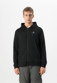 Joven frente a la cámara con una sudadera con cremallera negra y el logo de O’Neill en el pecho, y pantalones grises, de pie contra un fondo blanco liso.