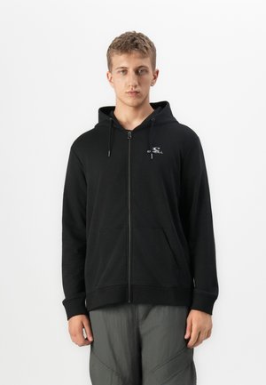 Un jeune homme regardant vers l'avant, portant un sweat à capuche zippé noir avec le logo O'Neill sur la poitrine et un pantalon gris, se tenant devant un fond blanc uni.