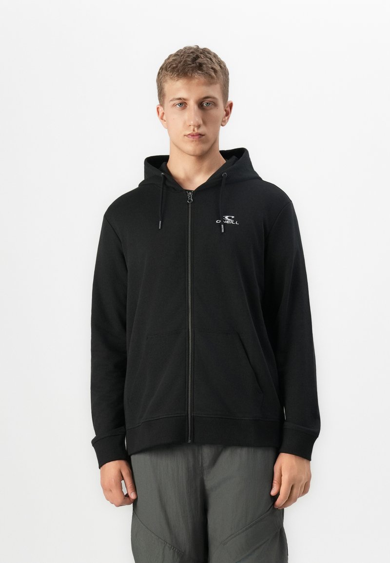 Joven frente a la cámara con una sudadera con cremallera negra y el logo de O’Neill en el pecho, y pantalones grises, de pie contra un fondo blanco liso.