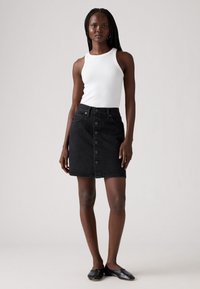 Falda de mezclilla negra con cierre de botones en la parte delantera, dos bolsillos frontales y una forma ligeramente acampanada. Combinada con una camiseta de tirantes blanca de canalé y bailarinas negras.
