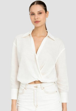 Donna dalla carnagione chiara e capelli castani indossa una camicetta avvolgente bianca con collo e maniche lunghe e pantaloni bianchi a vita alta, posando davanti a uno sfondo grigio.