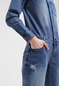 Jeansoverall i mörkblått, med långa ärmar, frontdragkedja, bröstficka och slitningar på benet. Slät textur.