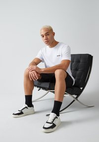Uomo con capelli biondi corti seduto su una moderna sedia in pelle nera, indossa una t-shirt bianca, pantaloni corti neri, calzini neri e scarpe nere e bianche.