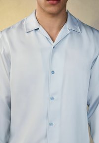 Camisa azul clara de manga corta en un tejido suave similar a la seda. Presenta un cuello clásico y cierre frontal con botones a juego.