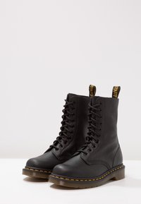 Botas de combate de cuero negro con 14 ojales, costuras amarillas y tiradores. Textura suave con una suela de goma gruesa.