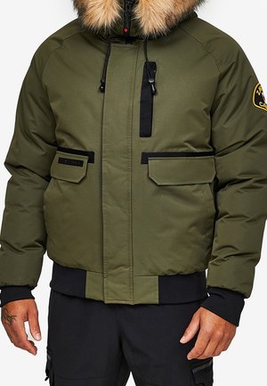 Oliivinvihreä talvitakki, jossa karvavuorattu huppu, musta vetoketju ja taskujen yksityiskohdat sekä merkki ylähihoissa, pidettynä mustujen housujen päällä.